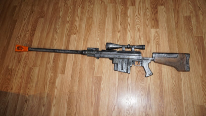 A large, black and silver toy gun with a scope and orange tip is lying on a wooden floor.